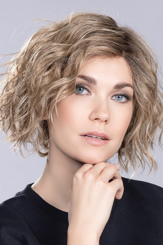 Model wearing a short length wig in blend of light honey blonde, light reddish brown and light brown with a darker root.