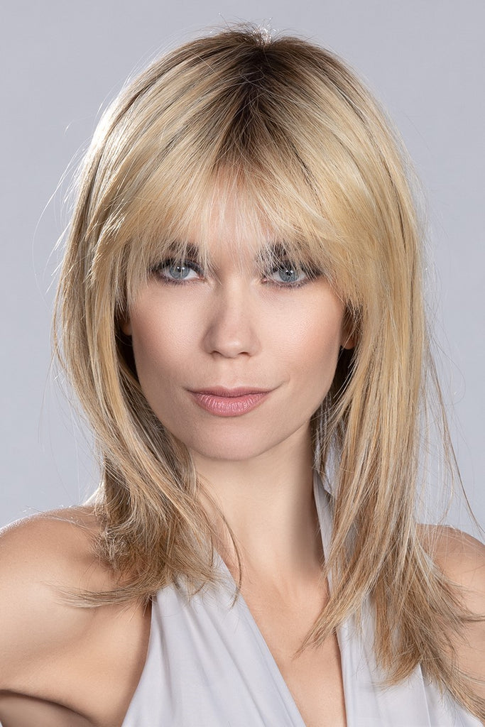 Women looking into the camera wearing a long length layered wig with wispy bangs. 