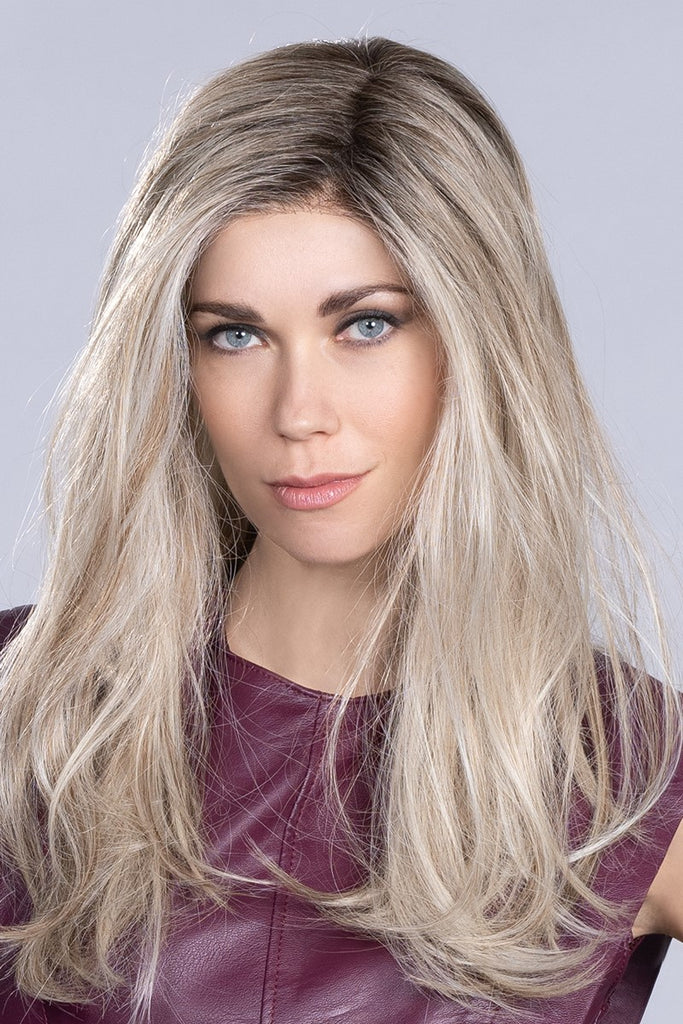 Women wearing a wig in a blend of cool blondes with a touch of honey blonde and a dark root.